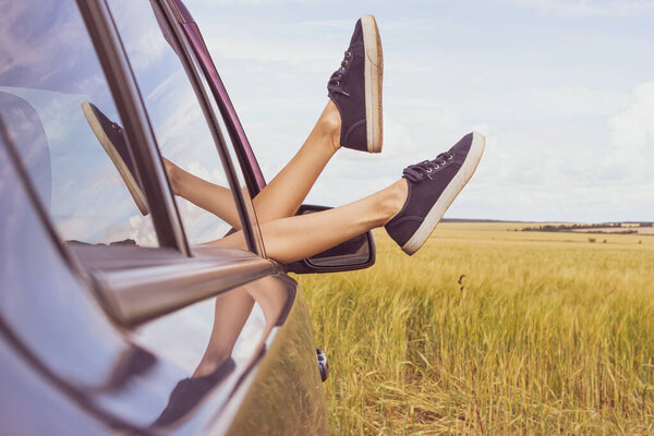 Women's legs in shabby blue sneakers stick out of the car window, against the backdrop of a wheat field. Cute funny image, car travel concept.