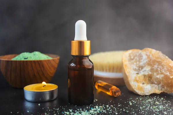 Glass bottle with pipette, candle, small bottle of essential oil, natural brush and decorative stone on dark background Steam - in air The concept of cosmetic procedures in bathroom, steam bath, sauna