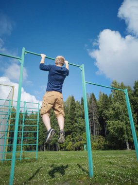 Genç Avrupalı sarışın adam ağaçların arka planında yatay bir barda asılı duruyor. Açık hava sporları konsepti. Adam spor ayakkabı, şort ve uzun kollu mavi bir tişört giyiyor..