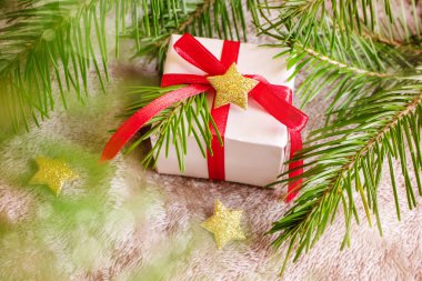 Gri ekoseli kırmızı kurdeleli, hafif ambalaj kağıdına sarılı küçük kare bir hediye. Yakınlarda altın yıldızlar, kırmızı bir Noel ağacı topu ve köknar dalları var. Yeni yıl konsepti, Noel, ev sıcacık tatil.