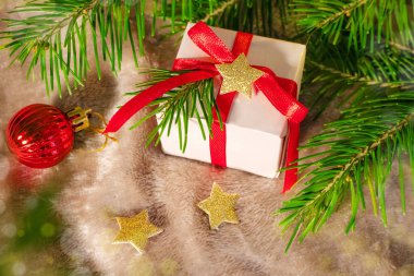 Gri ekoseli kırmızı kurdeleli, hafif ambalaj kağıdına sarılı küçük kare bir hediye. Yakınlarda altın yıldızlar, kırmızı bir Noel ağacı topu ve köknar dalları var. Yeni yıl konsepti, Noel, ev sıcacık tatil.