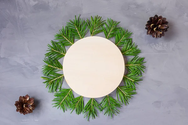 Spruce branches are laid out in form of circle, in the middle there is wooden round. Nearby is cone. New Years Christmas concept for postcard, banner. Gray background, copy space, minimalism, flat lay
