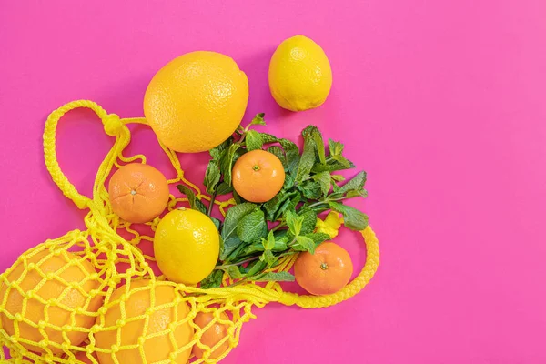 Oranges, tangerines, lemons in a yellow string bag. Citruses, vitamin C. Sustainability, zero waste, plastic free concept, vegetarianism, healthy food. Top view. Pink background.