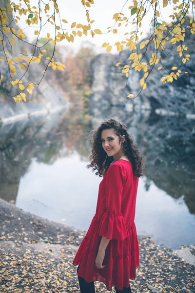 湖に立ち 笑みを浮かべて赤いドレスの美しい巻き毛の女の子 夢のような秋の背景 散歩でロマンチックな概念 — ストック写真