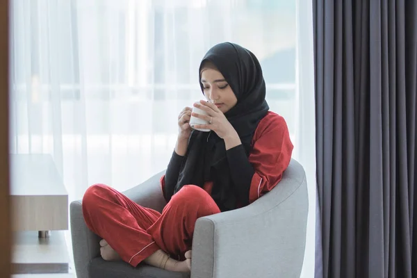 asian woman with scarf having a cup of coffee while sitting in a couch