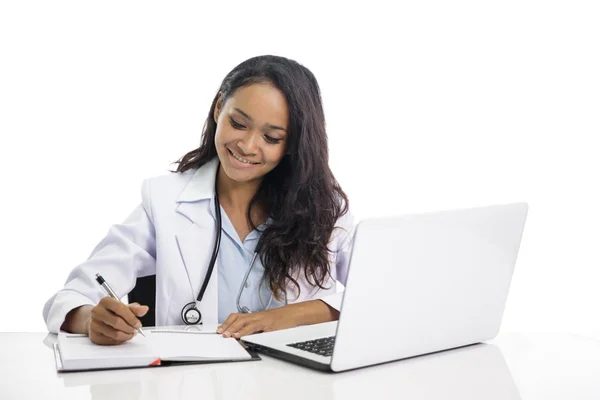 Schöne junge Ärztin arbeitet an ihrem Laptop und schreibt auf Notizen — Stockfoto