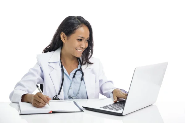 Bela jovem médico trabalhando em seu laptop e escrevendo em nota — Fotografia de Stock