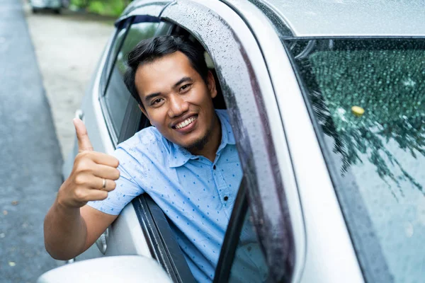 Молодой человек за рулем машины открывает окно, улыбается и показывает большой палец вверх — стоковое фото