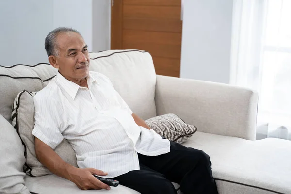 Retrato de un anciano solo sentado en el sofá sosteniendo el control remoto mientras — Foto de Stock