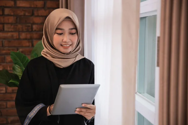 Jonge vrouw hijab vasthouden en kijkt een tablet staan in de buurt van ramen — Stockfoto