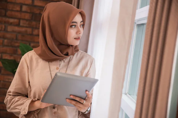 Wanita muda cantik terselubung memegang tablet dan melihat keluar jendela berdiri di dekat tirai — Stok Foto