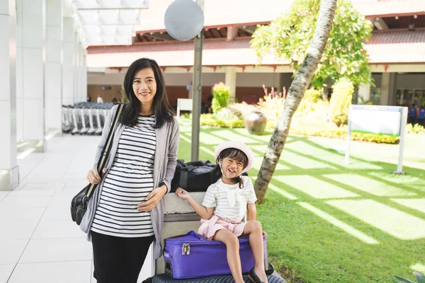 アジアの母親は、スーツケースの上に座っている娘の隣にバッグを運んで立っていた — ストック写真