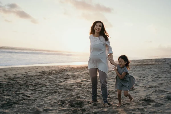 Junge schöne schwangere Frau mit ihrer kleinen süßen Tochter, die am Strand spielt — Stockfoto