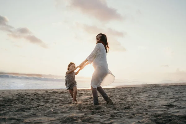 Jovem bela mulher grávida com sua filhinha fofa brincando na praia — Fotografia de Stock