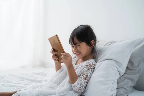 Kid liggande på sängen klädd i vitt med hjälp av mobiltelefon — Stockfoto