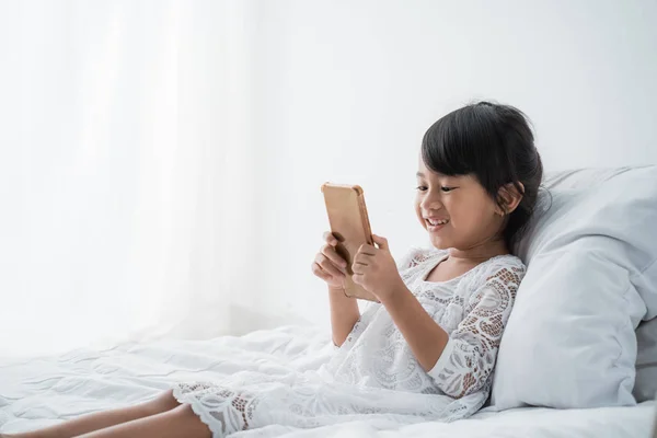 Kid liggande på sängen klädd i vitt med hjälp av mobiltelefon — Stockfoto