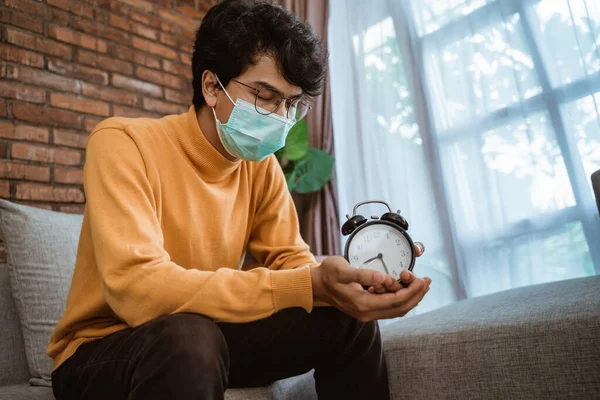 Homem vestindo uma máscara parece triste quando segurando um despertador — Fotografia de Stock