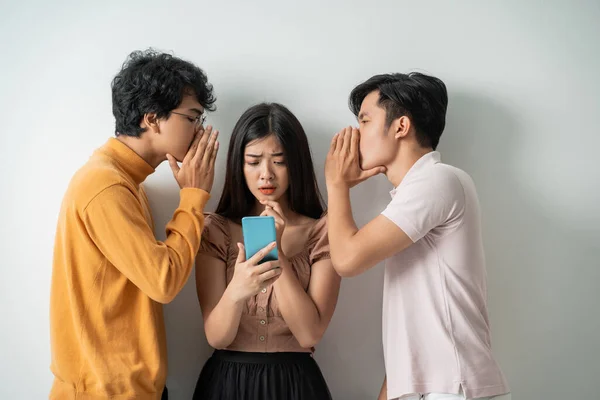 Two young men whispering to a young girl using a smart phone while standing — Stock Photo, Image