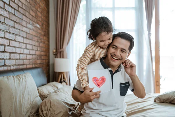 Asiático papá sonríe cuando él es abrazado por su pequeña hija por detrás mientras sostiene un papel con un corazón — Foto de Stock