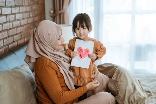 Gadis kecil tersenyum memegang kertas berbentuk hati sambil duduk di pangkuan ibunya — Stok Foto