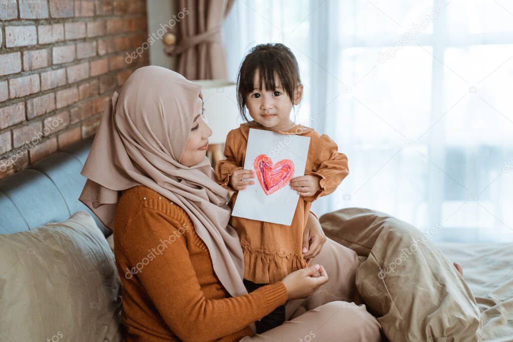 little girl smiles holding the heart-shaped paper while sitting on her mothers lap