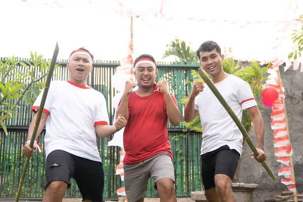 Sudut depan dari tiga pemuda dalam atribut merah dan putih dengan tangan terangkat dan memegang bambu berduri — Stok Foto