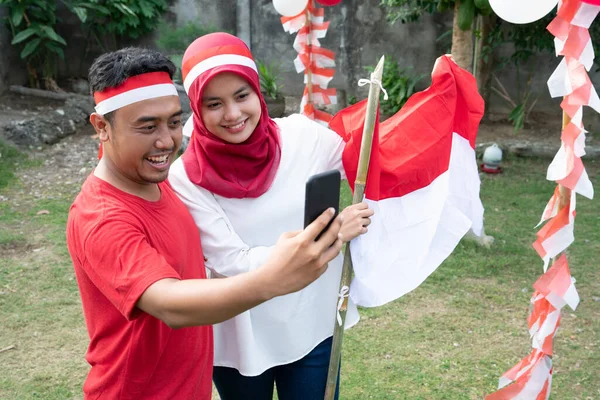Chłopiec i dziewczyna z hidżabem i czerwono-białe opaski stojące podczas robienia selfie razem trzymając bambusowe pałeczki z flagami Indonezji — Zdjęcie stockowe