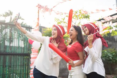 Kırmızı ve beyaz giysili üç neşeli kız cep telefonlarıyla selfie çekiyor.