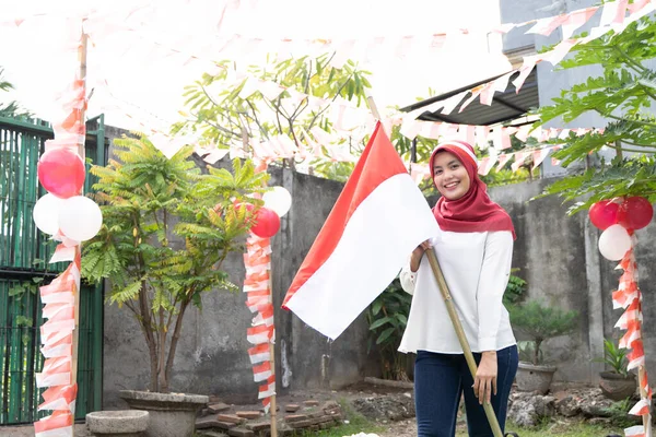 Девушка с хиджабом и красно-белой повязкой на голове держит бамбуковую палку с индонезийским флагом — стоковое фото