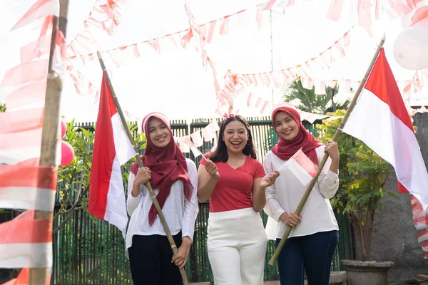 Tre asiatiska flickor bär röda och vita attribut leende bär indonesisk flagga bambu pinnar — Stockfoto