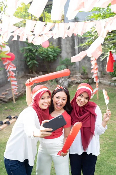 Tiga gadis ceria mengenakan pakaian merah dan putih mengambil selfie bersama-sama menggunakan ponsel — Stok Foto