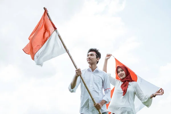 Ung man bär en vit skjorta hålla en pinne genom att höja den indonesiska flaggan bakom honom en ung kvinna som håller en flagga — Stockfoto