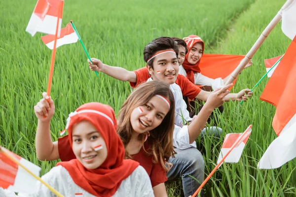 Hombres y mujeres jóvenes asiáticos sostienen y ondean la bandera indonesia mientras usan atributos rojos y blancos en los campos de arroz — Foto de Stock