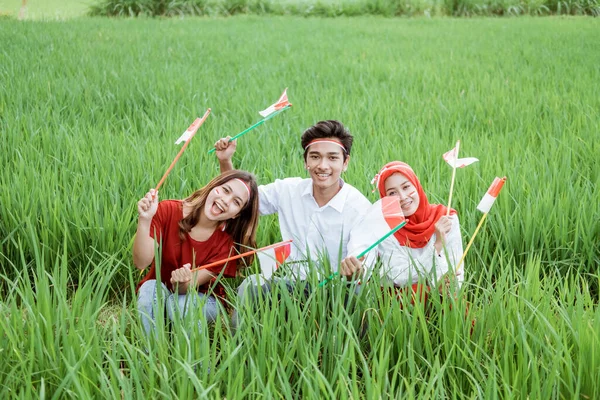 Jóvenes asiáticos sonríen sosteniendo pequeñas banderas rojas y blancas sentadas en medio de campos de arroz — Foto de Stock