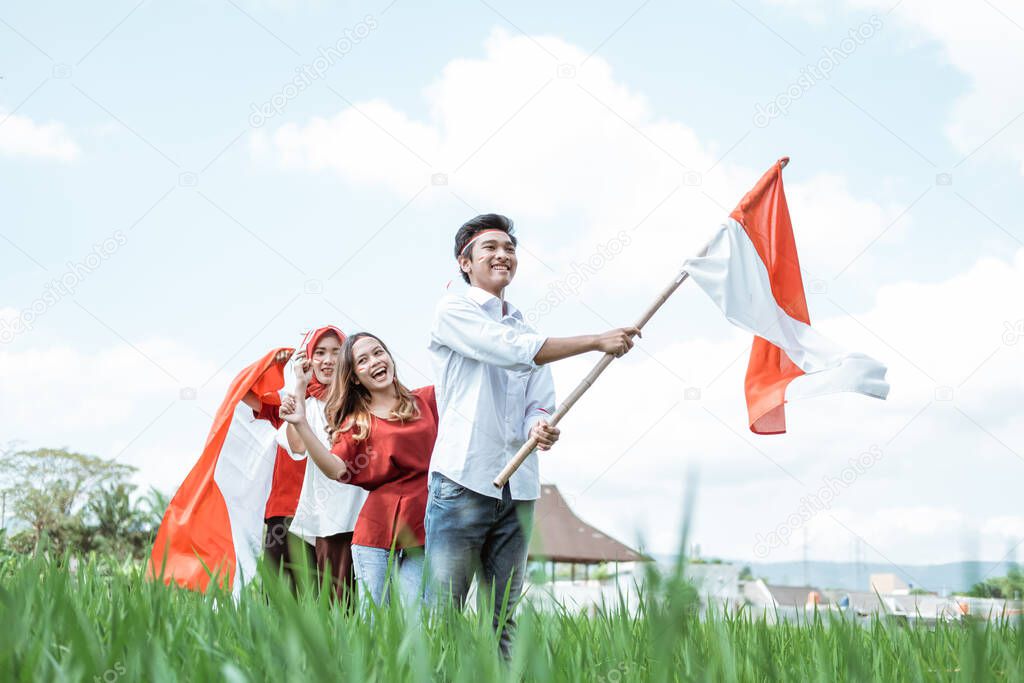 Asian man holds and raises Indonesian flag while following behind his friend wearing red and white attributes in the rice field