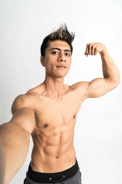 Joven con el cuerpo musculoso sosteniendo un teléfono inteligente para selfie con una mano muestra bíceps musculoso de pie mirando hacia adelante y mirar a una cámara — Foto de Stock