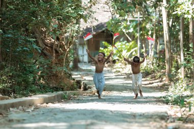 İki Asyalı çocuk kırmızı ve beyaz bayrak tutarken birbirlerinin peşinden koşuyorlar.