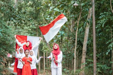 Bir grup Asyalı küçük kız, köy yolunda kırmızı ve beyaz bayrağı tutarken ve kaldırırken kahkahalarla gülüp şarkı söylüyorlar.