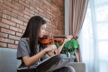 Kanepede keman tutan Asyalı kız keman çantasından kemanı alır ve keman yayı ile test eder.