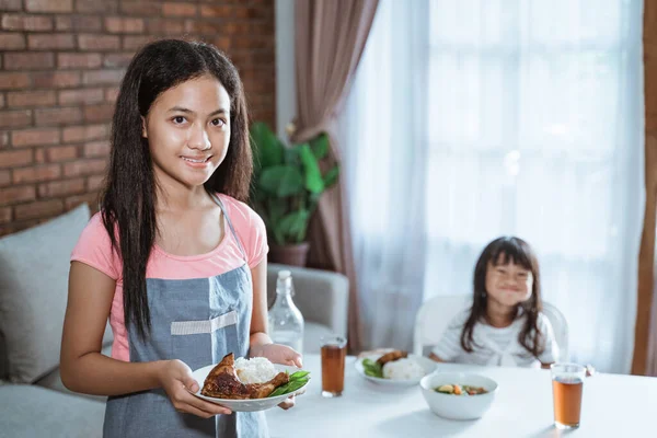 Tersenyum gadis remaja mengenakan celemek membawa sepiring makanan dan melihat kamera — Stok Foto