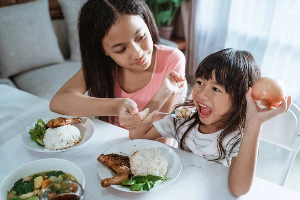 Asiático pequeña chica es excitado cuando su mayor hermana alimentar su mientras comer juntos en el comedor mesa — Foto de Stock