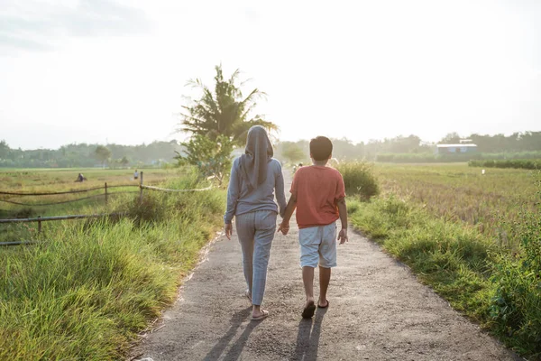Uitzicht van achteren gesluierde moeder en haar zoon lopen weg van de weg in het midden van rijstveld — Stockfoto