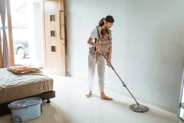 Asian Mom in white clothes with a baby in carrier baby mops the floor — Stock Photo, Image