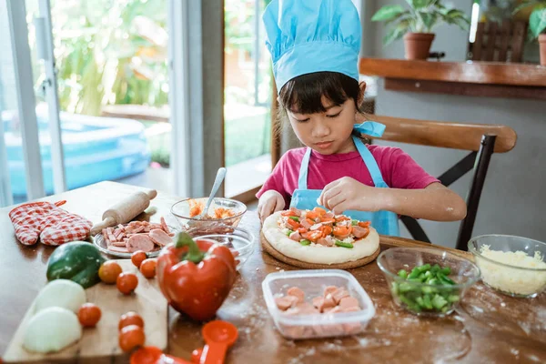 Asyalı şef şapkalı şirin küçük kız domatesli ve rendelenmiş peynirli pizza yapıyor. — Stok fotoğraf