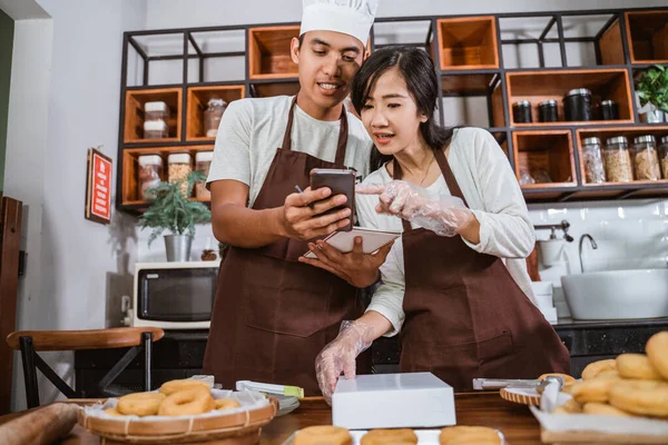 Asijský samec kuchař ukazuje smartphone na samici kuchař při přípravě koblihy objednávky — Stock fotografie