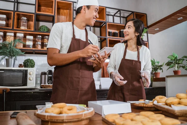 Chef masculino asiático sosteniendo el libro de pedidos mientras conversa con la chef femenina mientras prepara las órdenes de rosquillas —  Fotos de Stock