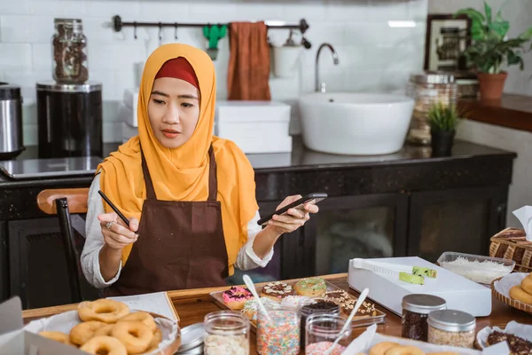 Eine verschleierte Frau trägt eine Schürze mit einem Handy und zählt die Anzahl der Donut-Bestellungen — Stockfoto