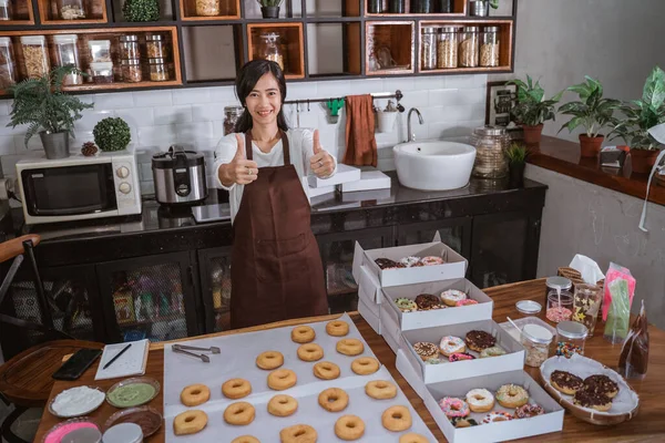 Asiática chef feminino sorrindo na cozinha moderna com os polegares para cima na frente de um donut em uma mesa — Fotografia de Stock