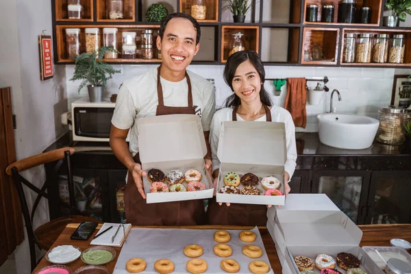 Sorrindo jovem casal na cozinha moderna de pé carregando caixas de donuts na frente de uma mesa — Fotografia de Stock