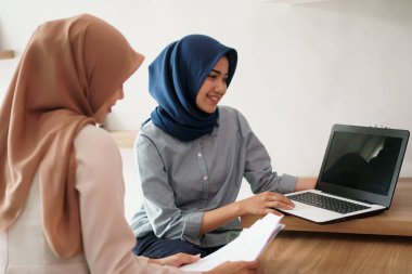 Çekici, genç Müslüman iş kadını laptopta çalışıyor ve masadaki modern ofisinde arkadaşıyla otururken gülümsüyor.
